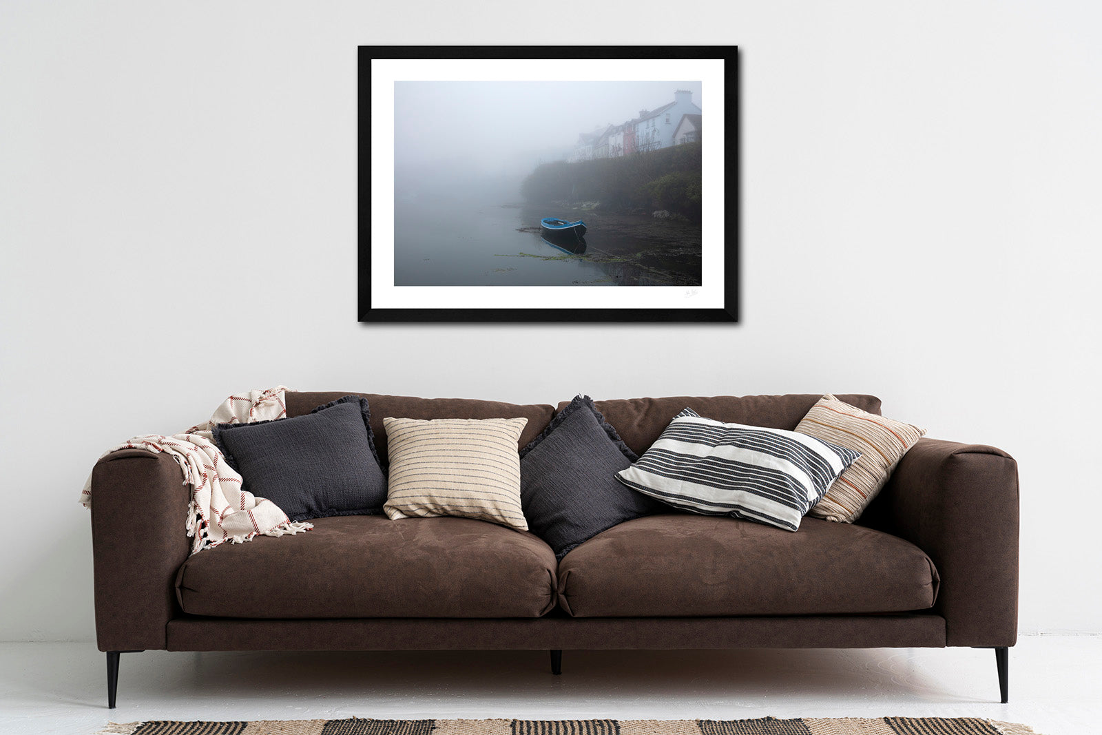 a framed fine art photograph of a rowing boat moored in Roundstone Harbour, Connemara in dense fog with only a few of the towns buildings visible hanging on a white wall above a big brown sofa 