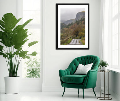 a framed fine art photograph of Glencar Valley in autumn with stunning forests and steep mountain cliffs hanging on a white wall above a green chair in a minimalist room 