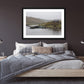 a framed fine art photograph of a fishing hut and row boat mooring on the Owenmore River on a rainy day with a few grazing sheep hanging on a dark bedroom wall above a wooden double bed