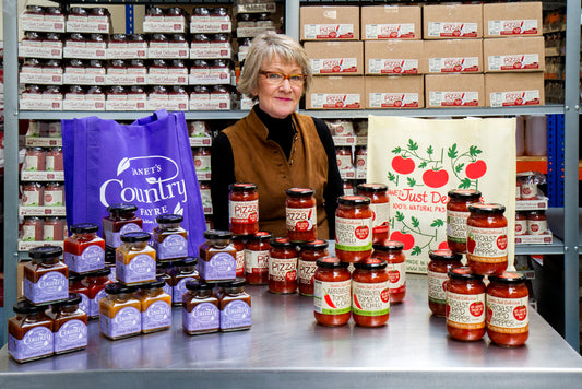 Portrait of Janet sitting behind a table with all her chutneys and sausces displayed