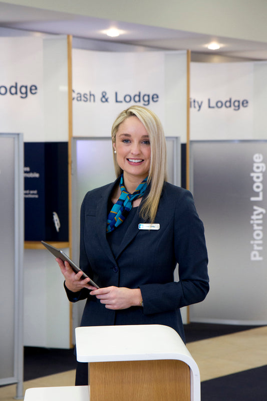 a uniformed young woman stands to welcomes customers into the self service area of a bank 